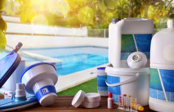 Swimming pool service equipment with chemical cleaning products and water testing tools on wood table by a pool.